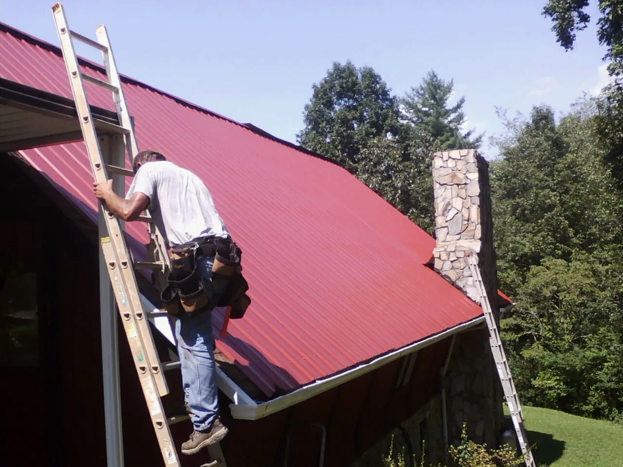 Roofer on a ladder beside a red metal roof and stone chimney — Soffit & Fascia Repair by Roofing Brothers in Fall Branch